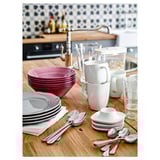 Comptoir de cuisine avec des tasses VÄRDERA blanches empilées, des bols roses, des assiettes grises et des ustensiles en argent contre un mur carrelé.