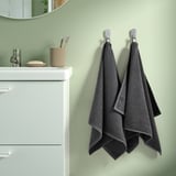 Deux serviettes grises VÅGSJÖN sont accrochées à des crochets muraux à côté d’une armoire blanche avec un porte-brosse à dents beige et un petit porte-savon, sur un mur vert pâle.