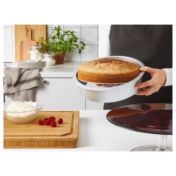Une personne tenant un gâteau dans un moule rond à base amovible, avec des framboises et de la crème sur une planche à découper, dans un environnement de cuisine.