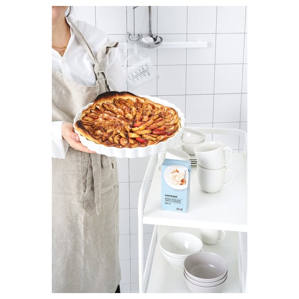 Une personne tient une tarte aux fruits sur une assiette blanche, à côté de tasses, de bols et d’une boîte sur laquelle on peut lire VARDAGEN.