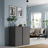Armoire VIHALS grise dans un salon moderne avec des murs bleus et des images encadrées en noir et blanc, avec des portes coulissantes et des objets décoratifs sur le dessus.