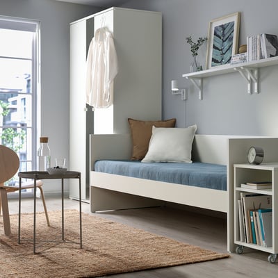 Chambre minimaliste avec lit de repos VIHALS blanc, coussins bleus, unités de rangement assorties, chaise en bois et grande fenêtre.