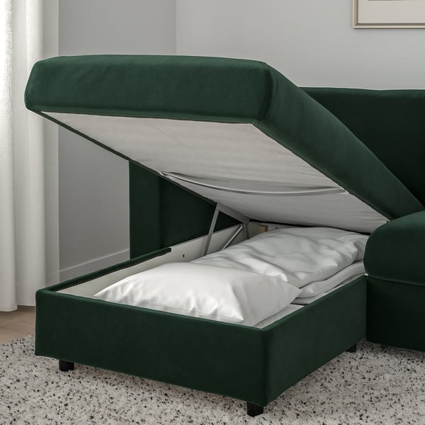 Canapé vert foncé avec un compartiment de rangement surélevé et accessible sous le siège, montrant les oreillers à l’intérieur.