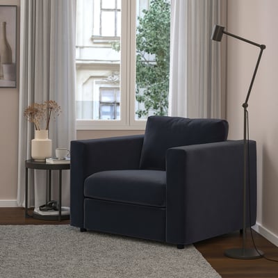 Salon moderne avec fauteuil VIMLE bleu marine, lampe, table d’appoint, rideaux gris, parquet et tapis.