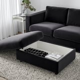 Repose-pieds VIMLE gris foncé avec rangement ouvert, rangement de livres et télécommande, à côté du canapé assorti.
