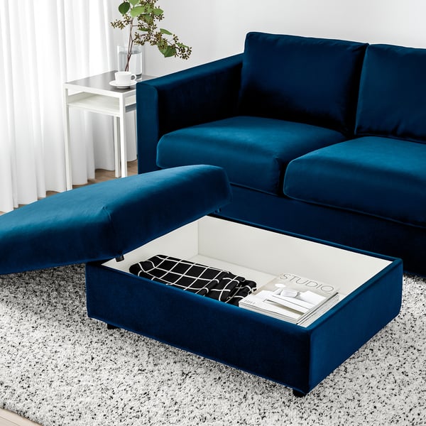 Canapé en velours bleu avec pouf ; Ottoman ouvert, révélant un espace de rangement avec des magazines à l’intérieur.