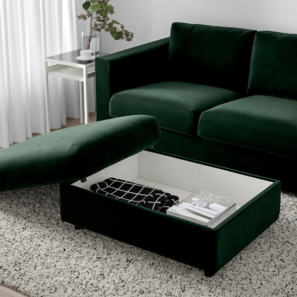 Salon moderne avec canapé sectionnel VIMLE vert, repose-pieds de rangement contenant des magazines et une télécommande.