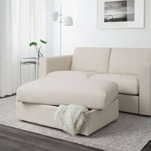 Salon moderne avec canapé sectionnel VIMLE beige doté d’un pouf avec rangement, surmonté d’une couverture blanche à franges.