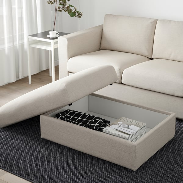 Canapé sectionnel VIMLE beige avec pouf de rangement ouvert, rempli de magazines sous un tapis gris.