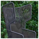 Fauteuil à oreilles en osier foncé avec coussin, posé à l’extérieur au milieu d’un feuillage vert.
