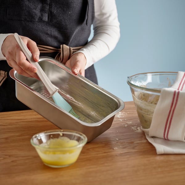 La personne graisse le moule à pain avec une spatule bleue, préparant la cuisson du pain. L’étain repose sur une table en bois avec un bol de beurre fondu.
