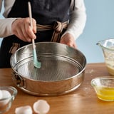 Une personne utilisant un moule à charnière, l’enrobant d’huile pour la cuisson. La poêle est posée sur une table entourée d’ingrédients.