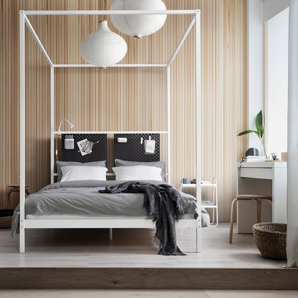 Chambre moderne avec lit à baldaquin blanc VITARNA, draps gris, deux lampes sphériques, mur en panneaux de bois, commode blanche avec miroir, plante et lampe.