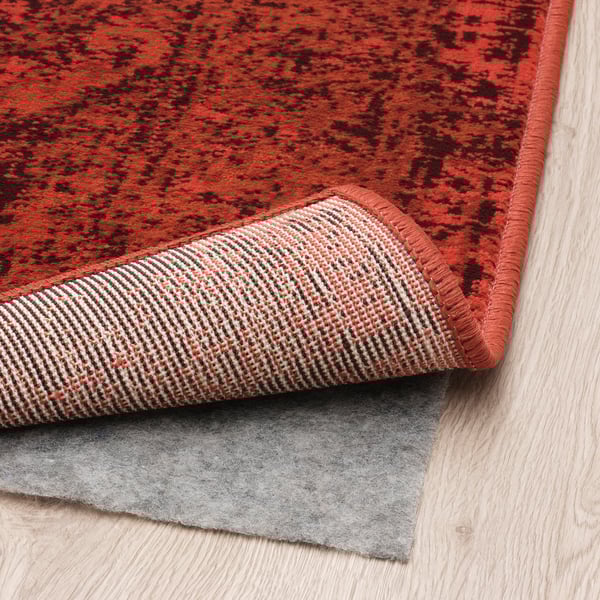 Tapis à motifs rouges à poils ras, adapté pour les salles à manger ou les salons, illustré avec un tapis antidérapant gris en dessous.