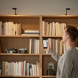 Un homme ajuste les lumières de son smartphone vers une étagère en bois remplie de livres et d’appareils photo.