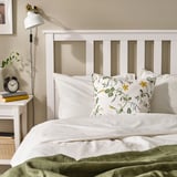 Gezellige slaapkamer met wit bed, groene deken, bloemenkussen. Wandklok links, witte wandlamp.