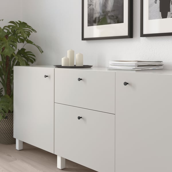 Witte kast met zwarte knoppen; Boven houdt kaarsen, onder heeft lades, tegen muur met ingelijste kunst.