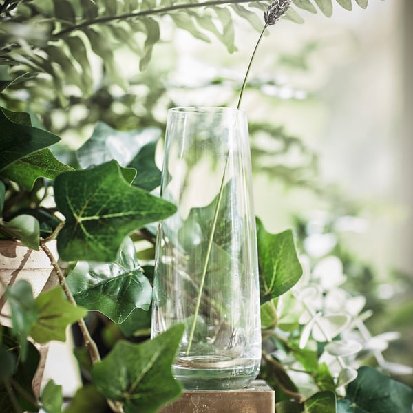 Heldere glazen vaas met een enkele witte bloem, omgeven door groene bladeren.
