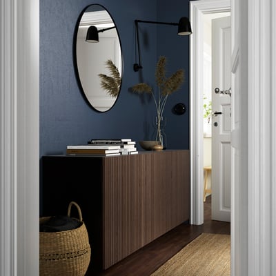 Moderne hal met BESTÅ dressoir, blauwe muur, ronde spiegel, wandlampen, boeken, mand, vaas met lange planten. Deur open naar lichte kamer.