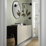 Wit BESTÅ dressoir met spiegel, boeken, vazen, planten; Groene muur, zwarte lampen, gevlochten mand.