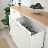 Witte kast met open lade, gaasmand binnenin, houten aanrechtblad met schaal met donker fruit, potplant in de buurt.