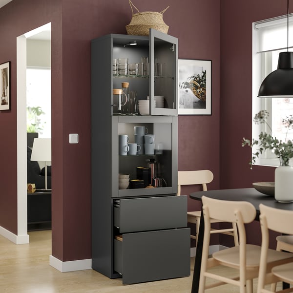 Moderne donkergrijze BESTÅ kast met glazen deuren en lades, met keukengerei tegen een bordeauxrode muur.