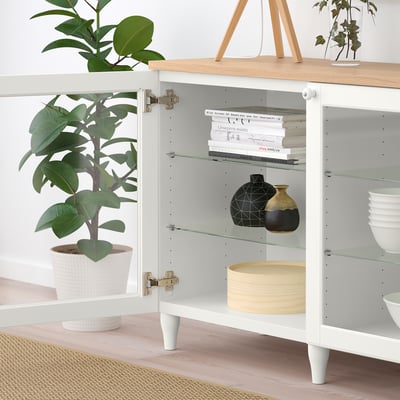 De foto toont een wit BESTÅ dressoir met glazen deuren, met planken met boeken, vazen, schalen en potplanten op een houten vloer.