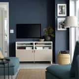 Moderne woonkamer met BESTÅ tv-meubel, flatscreen, bloemen, foto's, mand, lamp. Muur is donkerblauw.