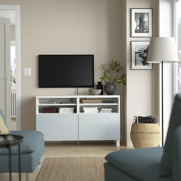 Moderne woonkamer met BESTÅ tv-meubel, blauwe fauteuils, flatscreen-tv, boeken en inrichting. Beige muur met foto's, vloerlamp en gevlochten mand.