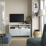 Moderne woonkamer met BESTÅ tv-meubel, blauwe fauteuils, flatscreen-tv, boeken en inrichting. Beige muur met foto's, vloerlamp en gevlochten mand.