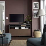 Moderne woonkamer met grote flatscreen-tv op BESTÅ houten standaard. Paarse muren, ingelijste foto's, blauwe stoel, rieten mand en lamp.