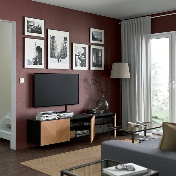Kastanjebruine woonkamer met aan de muur gemonteerd BESTÅ tv-meubel, kabels zichtbaar, planken met boeken en vaas, glazen salontafel voor grijze bank.