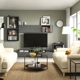 Moderne woonkamer met saliegroene muren, witte banken, glazen salontafel en zwart entertainmentcentrum met tv, boeken en decoraties.