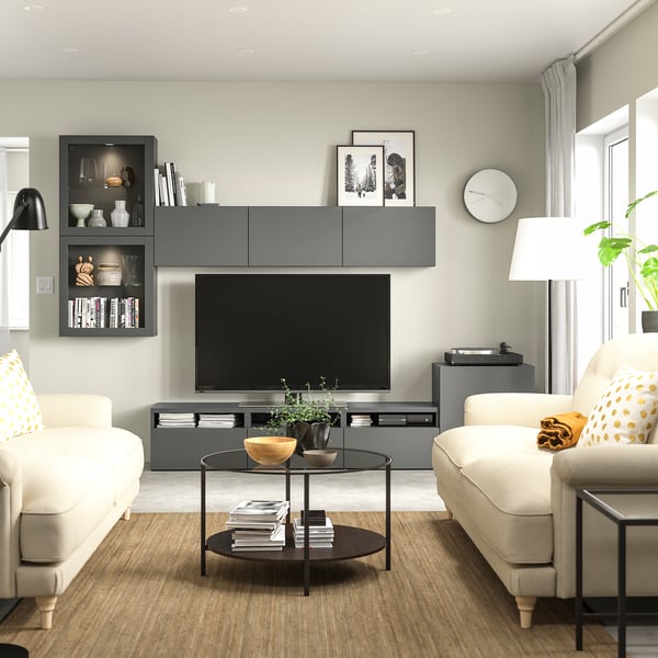 Moderne woonkamer met een grijs BESTÅ tv-meubel, witte banken met gele kussens, een glazen salontafel en een zwarte staande lamp.