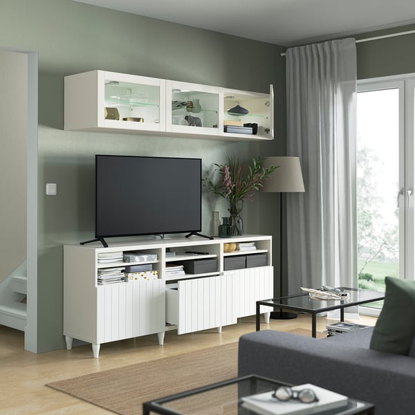 Moderne woonkamer met wit BESTÅ tv-meubel, flatscreen-tv, wandkasten, grijze bank, salontafel en groot raam met gordijnen.