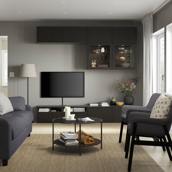 Moderne woonkamer met BESTÅ tv-meubel, grijze bank, kleurrijke kussens, glazen salontafel, lamp en groot raam.