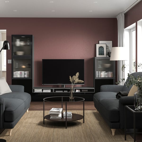 Moderne woonkamer met kastanjebruine muren, voorzien van een zwart BESTÅ tv-meubel, grijze banken en een ronde salontafel met boeken en een plant.