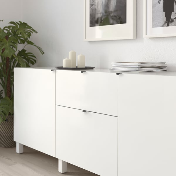 Witte opbergkast met moderne handgrepen, kaarsen en boeken tegen een muur met ingelijste foto's en een plant.
