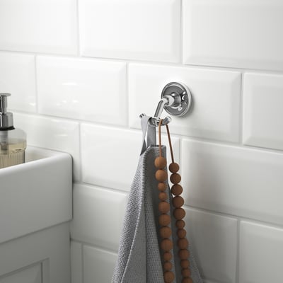 Een chromen dubbele haak aan een betegelde badkamermuur bevat een grijze handdoek en een houten kralensnoer, in de buurt van een witte gootsteen met zeepdispenser.