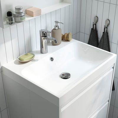 Kleine, moderne wastafel in de badkamer met chromen kraan en zeepdispenser. Chromen handgrepen en afvoer. Betegelde, witte muurachtergrond met opslag en handdoeken.
