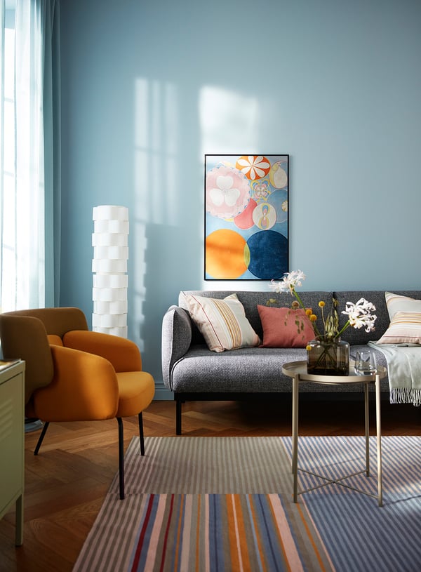 Lichte woonkamer met blauwe muren, grijze bank, gele stoel, kleurrijk gestreept vloerkleed, moderne kunst, hoge witte lamp en bloemen op salontafel.