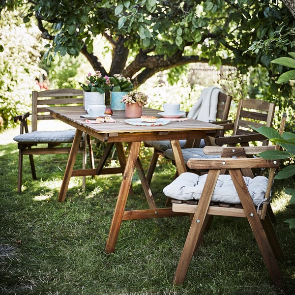 Buiten patio set met houten tafel en stoelen onder een boom. Tafel heeft borden, kopjes en potplanten. Stoelen hebben grijze kussens.