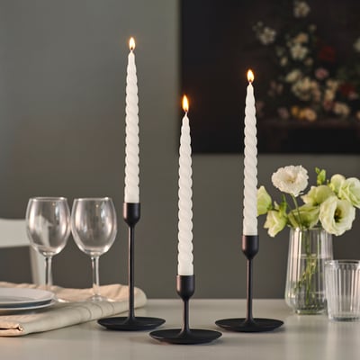 Drie brandende spiraalkaarsen in zwarte houders op een tafel met glazen en bloemen.