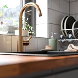 Moderne keuken met messing DELSJÖN kraan met hoge uitloop, bij de vaat, zeepdispenser en plant, tegen betegelde muur.