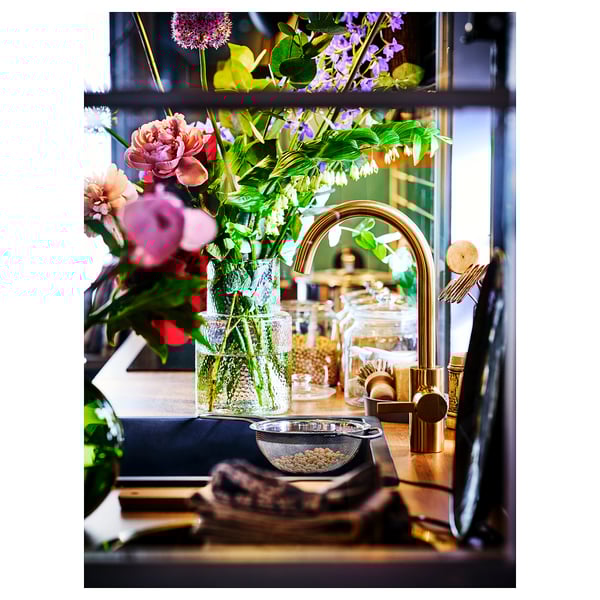Een gootsteen met een goudkleurige DELSJÖN kraan. Boeket bloemen, houten snijplank en keukenartikelen netjes gerangschikt.
