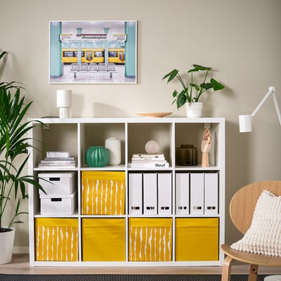 Moderne leefruimte met witte rekken met daarin gele en witte stoffen dozen, decoratieve artikelen en planten. Kunstwerk hangt erboven.