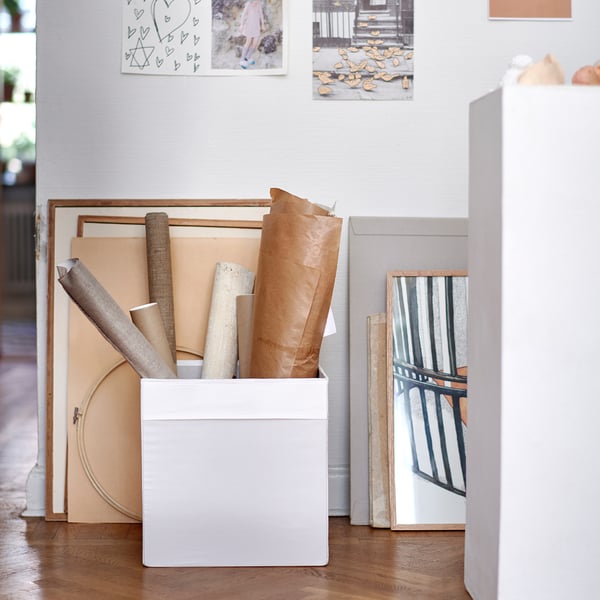 Witte stoffen opbergdoos met opgerolde papieren, geplaatst op houten vloer tegen een lichte muur met foto's.
