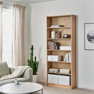 Gezellige woonkamer met lichte meubels. Stoel, kopje, hoge boekenkast, cactus, kunst aan de muur.