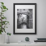 Aan een muur hangt een zwart-witte zwart-witte brug en gracht. Op een plank eronder staan boeken, een plant, een vaas en een rond voorwerp.