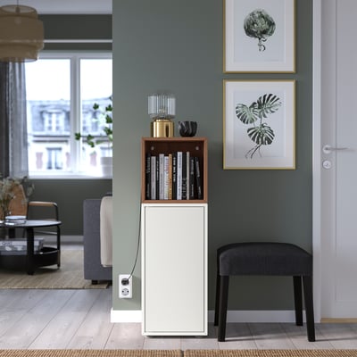 Moderne woonkamer met EKET witte kast, zwarte kruk, groene muur, ingelijste plantenprints, blauwe bank en houten vloer.